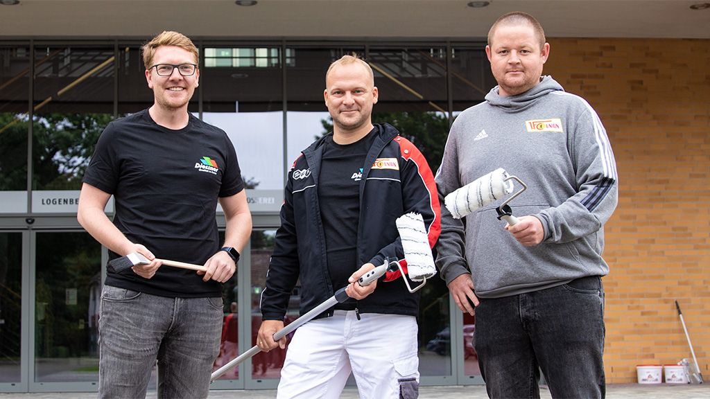 Drei Männer stehen vor einem Eingang. Sie halten Farbpinsel, einen Malerroller und einen Farbroller, gekleidet in T-Shirts mit Logos.