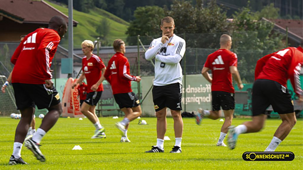Ein Trainer beobachtet eine Gruppe von Spielern bei einem Fußballtraining auf einem grünen Spielfeld.