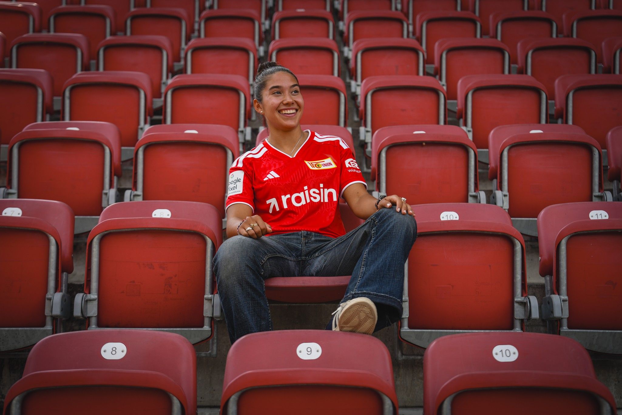 Eine lächelnde Person trägt ein rotes Trikot in einem leeren Stadion mit roten Sitzen.