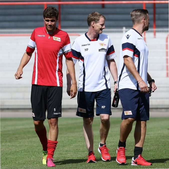 Drei Männer in Sportkleidung stehen auf einem Fußballfeld und unterhalten sich.