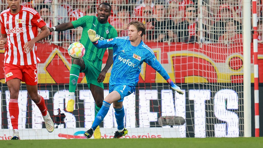 Goalkeeper in blue gear reaches for the ball while a player in a green jersey challenges him. Audience in the background.