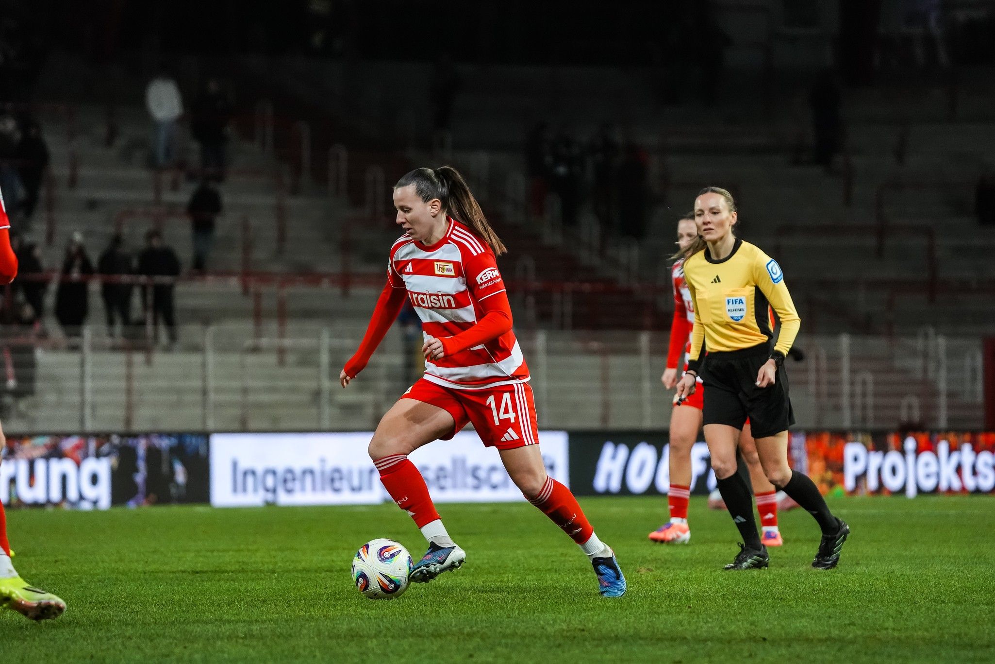 Spielerin in rotem Trikot dribbelt den Ball auf dem Fußballfeld, während eine Schiedsrichterin zusieht.