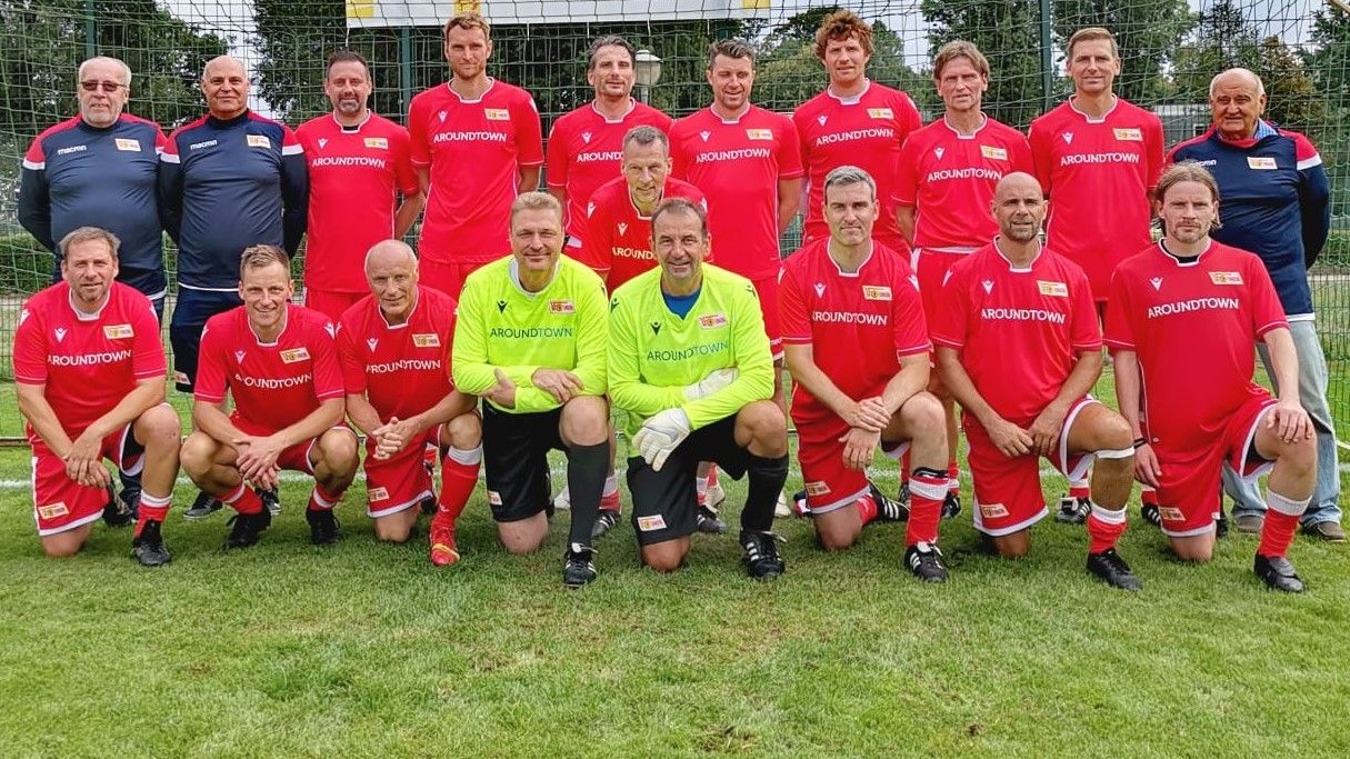 Eine Fußballmannschaft in roten Trikots posiert auf einem Rasenplatz. Im Hintergrund steht ein Tor.