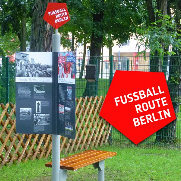 Schautafel zur Fußballroute Berlin, auf einem Pfosten montiert, mit einer Holzbank und einem Zaun im Hintergrund.