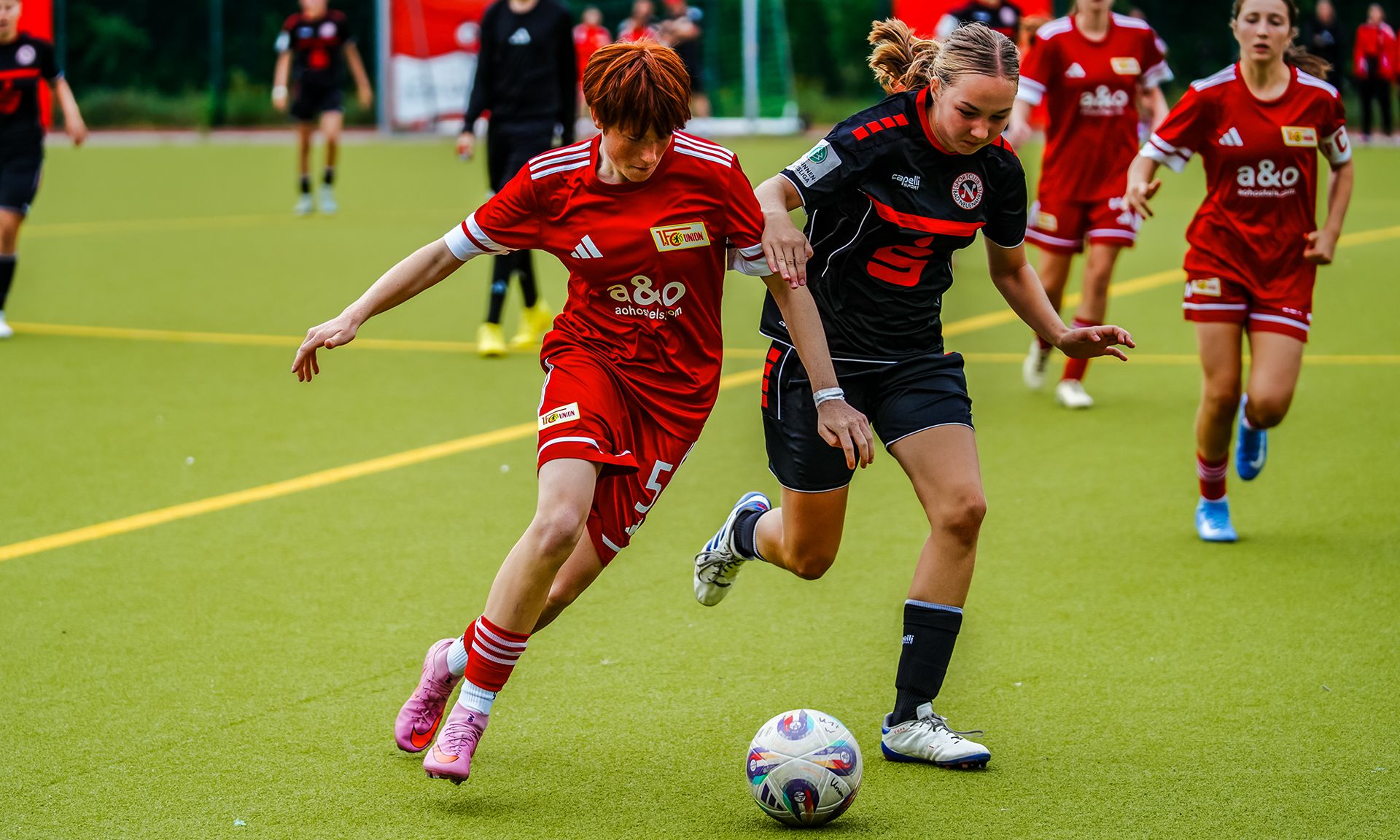 Zwei Fußballspielerinnen kämpfen um den Ball auf einem Kunstrasenfeld während eines Spiels.