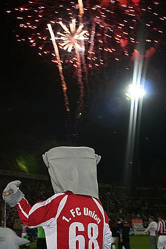 Ein Maskottchen mit einem Eimer auf dem Kopf trägt ein Trikot des 1. FC Union, während Feuerwerk am Nachthimmel explodiert.