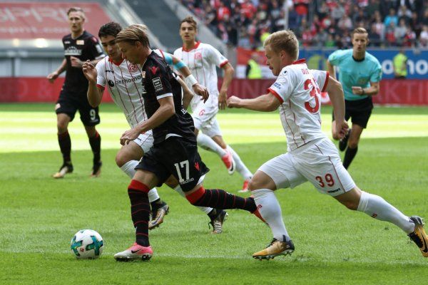 Game scene with players in black-red and white competing for the ball. Stadium atmosphere in the background.