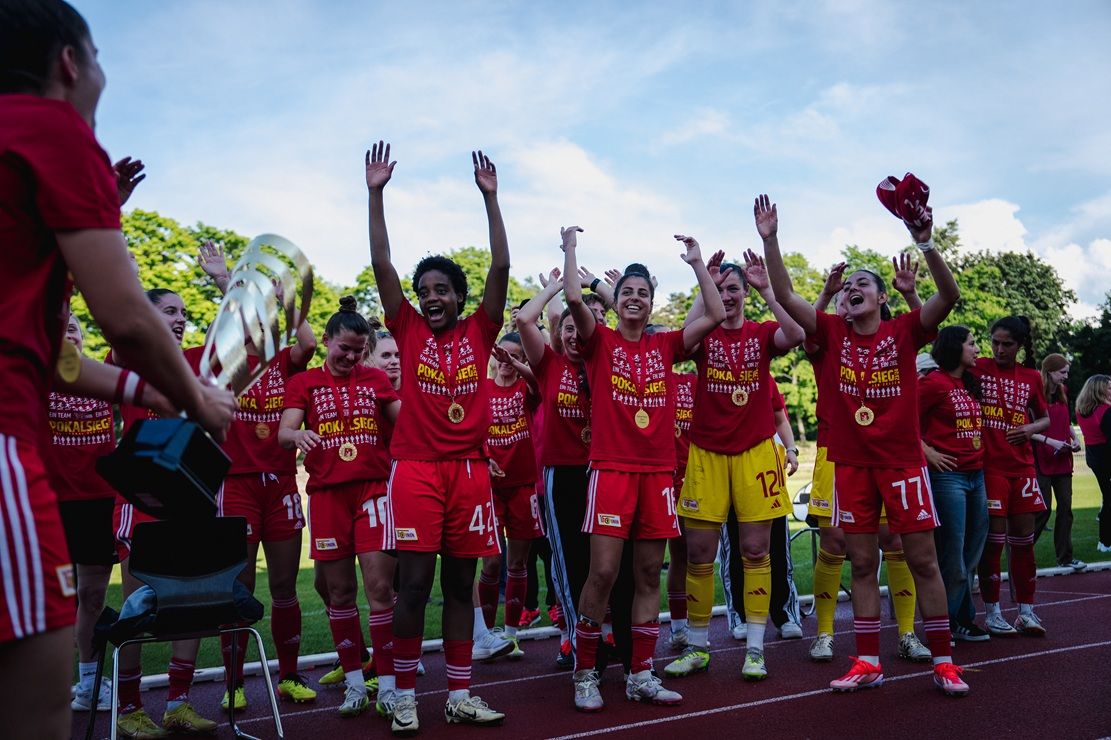 Frauenfußballteam in roten Trikots feiert mit Händen in der Luft und Trophäe auf dem Podium nach einem Sieg.