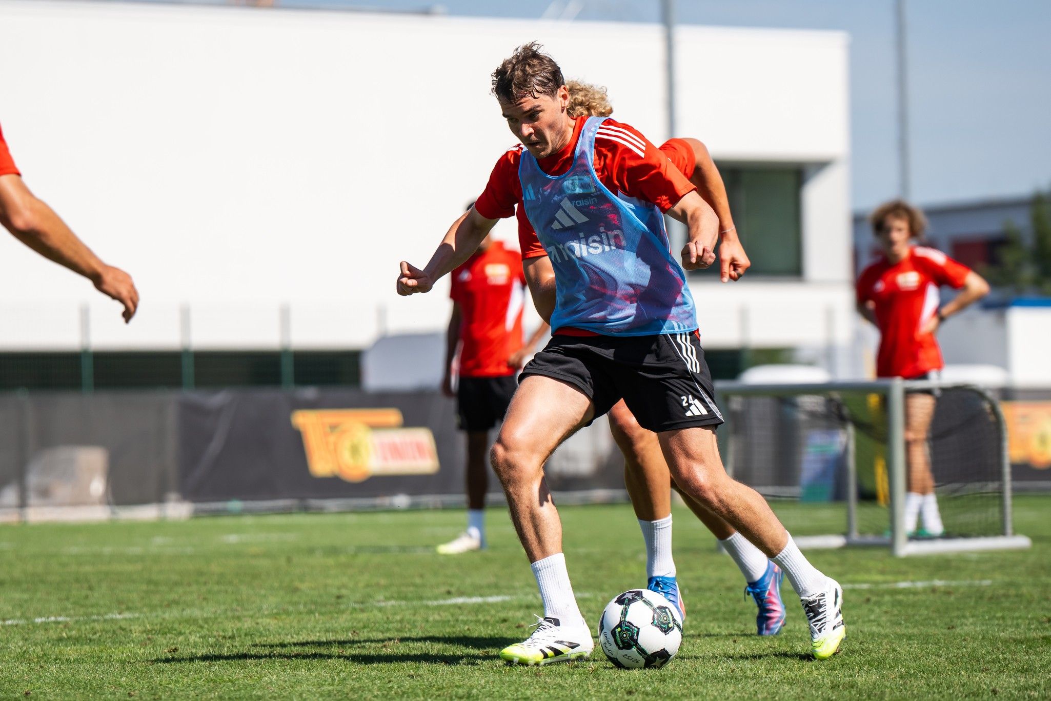 Spieler in rot-blauem Trikot dribbelt den Ball während eines Fußballtrainings auf dem Rasen. Hintergrund zeigt weitere Teammitglieder.