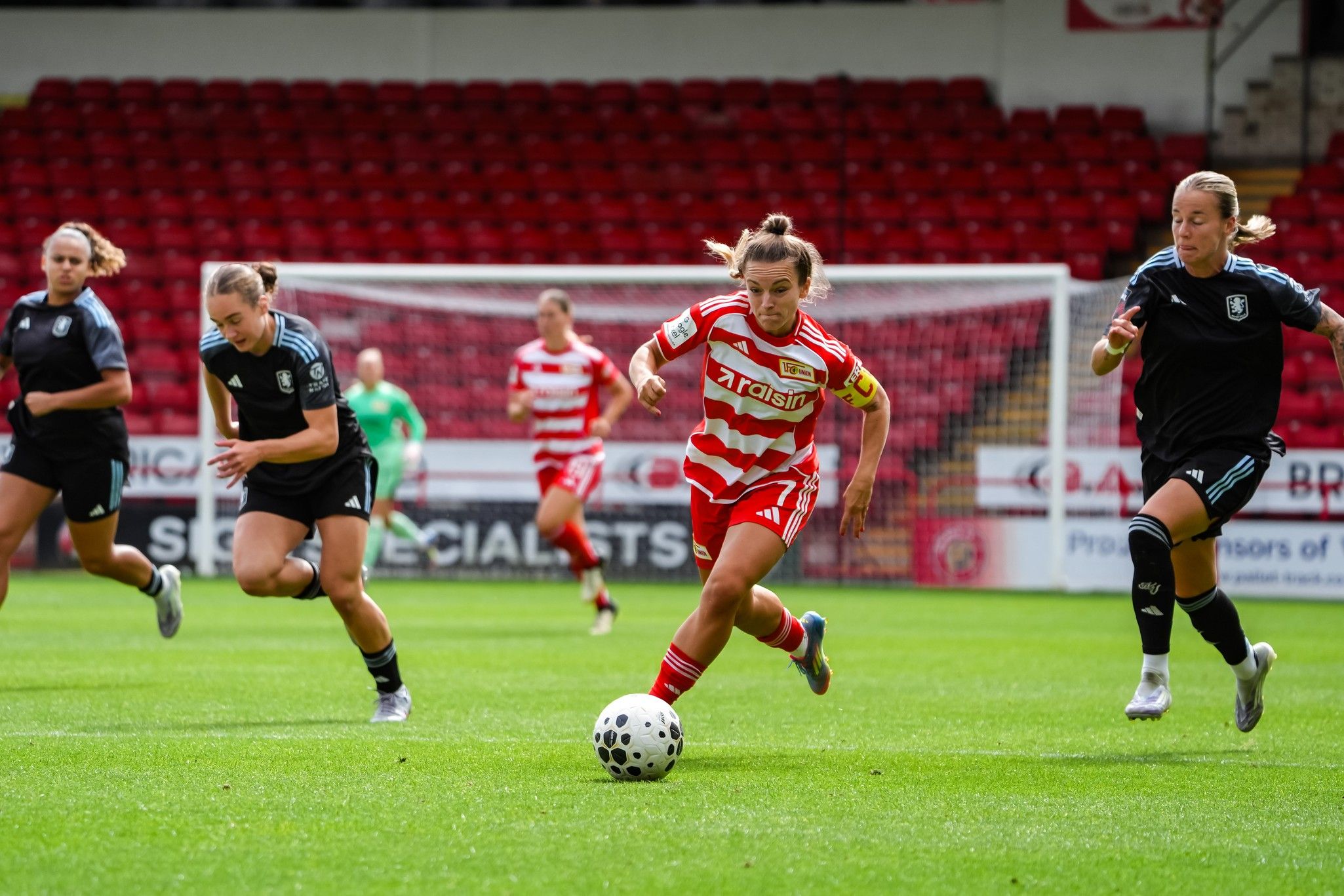 Eine Spielerin in rot-weiß gestreiftem Trikot dribbelt den Ball, während zwei Gegnerinnen auf dem Fußballplatz verfolgen.