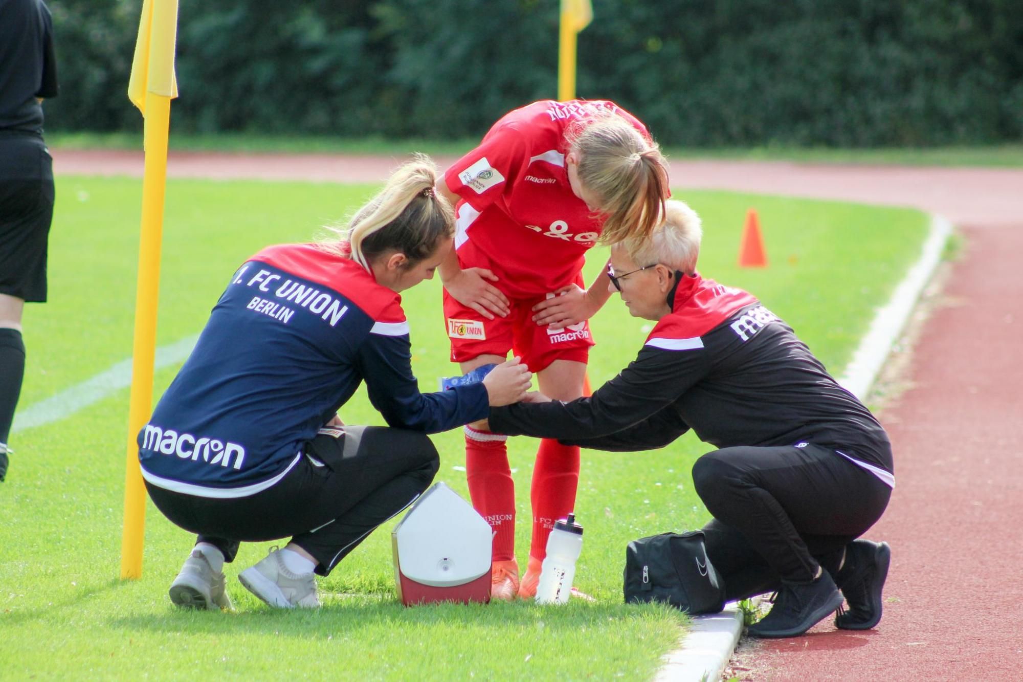 Zwei Betreuer stehen neben einer verletzten Spielerin, die sich auf das Spielfeld beugt. Sonne scheint, Fußballplatz im Hintergrund.