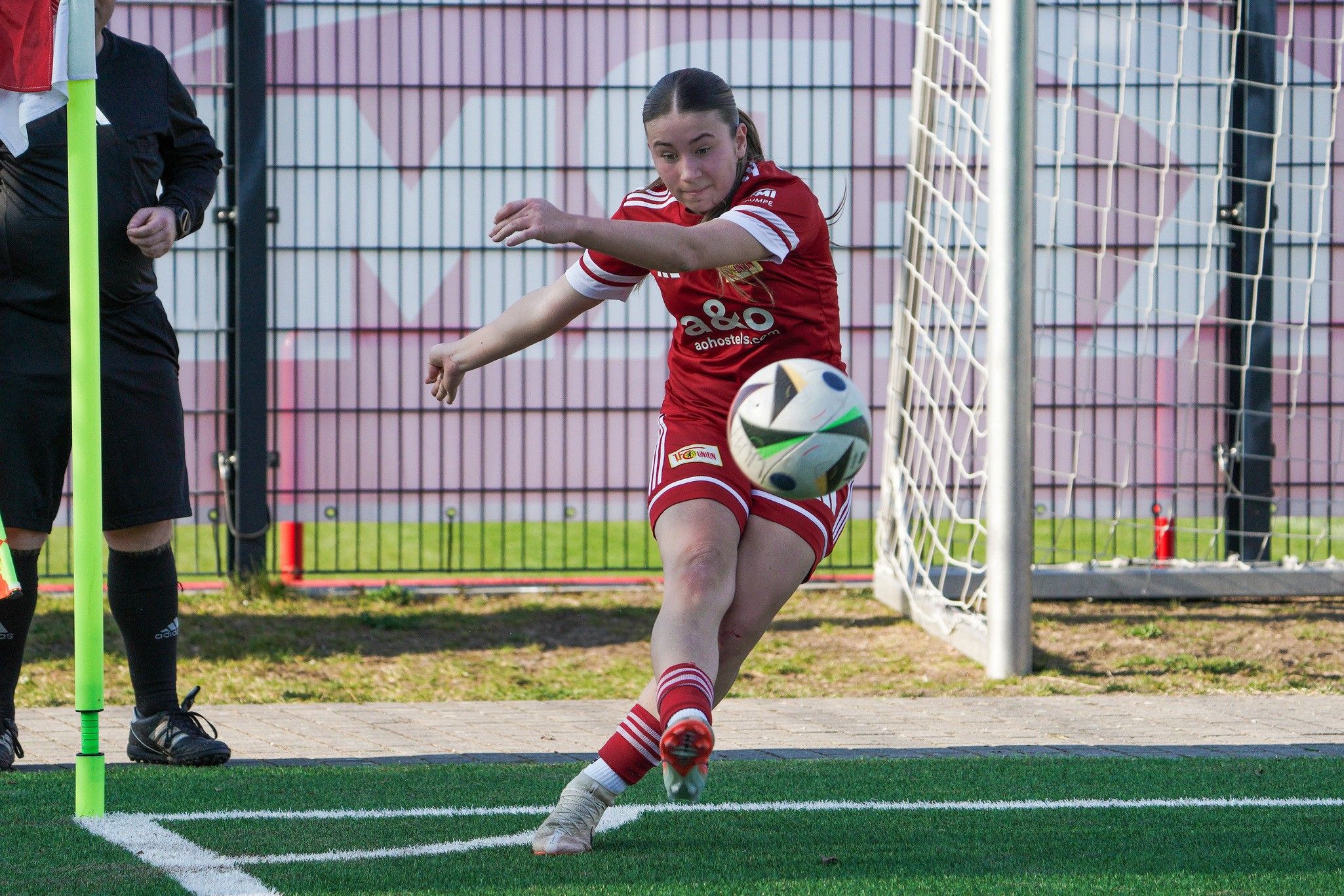 Spielerin in roter Fußballausrüstung führt einen Eckball aus, während ein Schiedsrichter im Hintergrund steht.