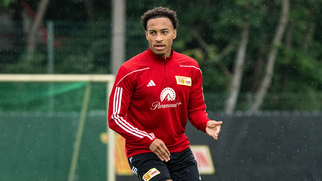 Ein Fußballspieler in rotem Trainingsanzug übt auf dem Platz bei Regen.