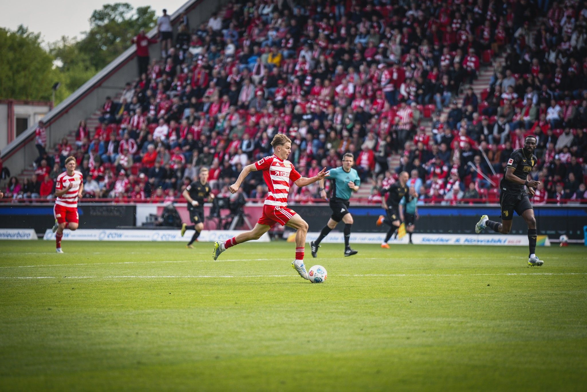 Ein Fußballspieler in roter und weißer Kleidung sprintet über das Feld, während Zuschauer im Hintergrund jubeln.