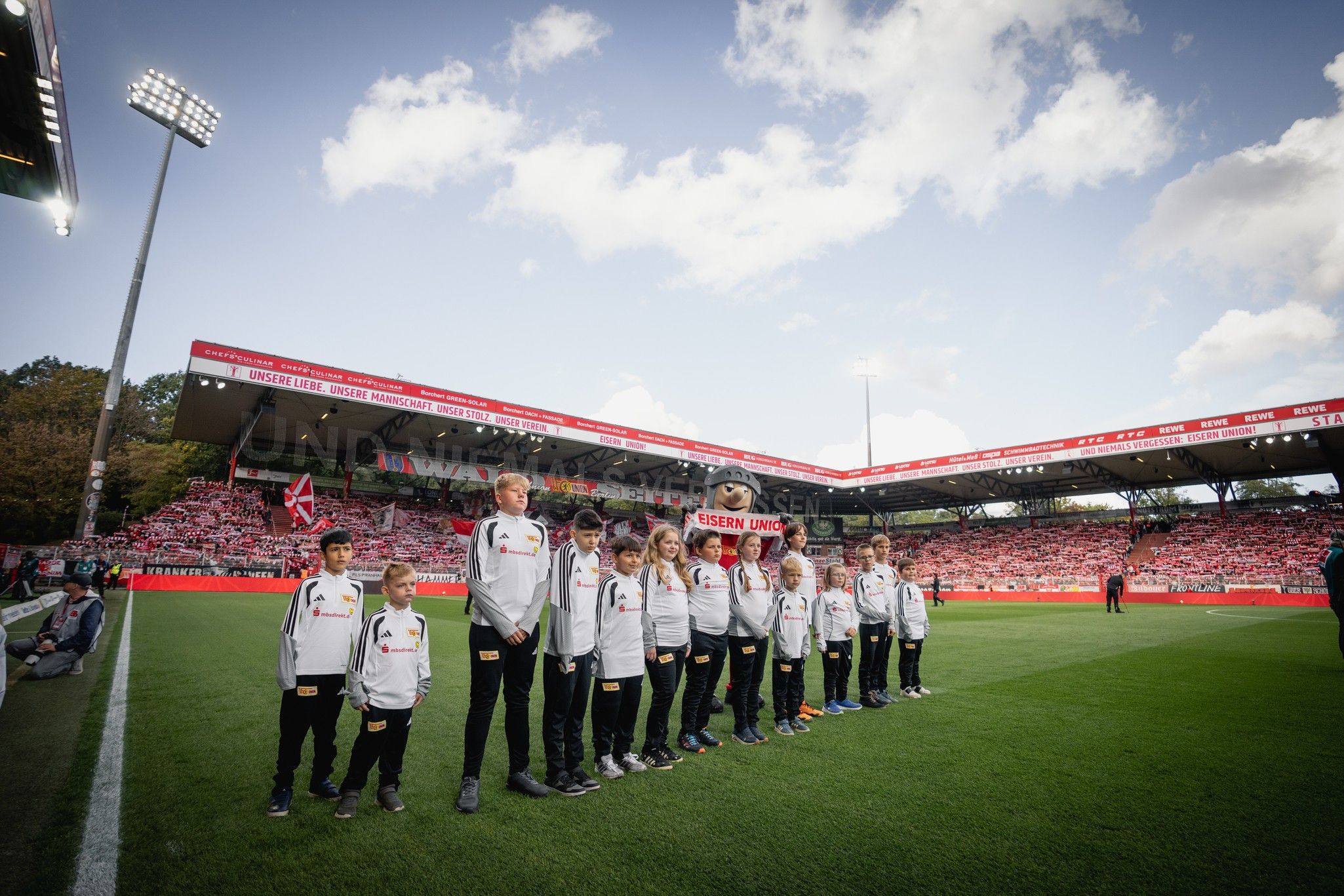 Kinder in Teamkleidung stehen in einer Reihe auf dem Spielfeld, während ein Maskottchen neben ihnen steht. Im Hintergrund ist das Stadion sichtbar.