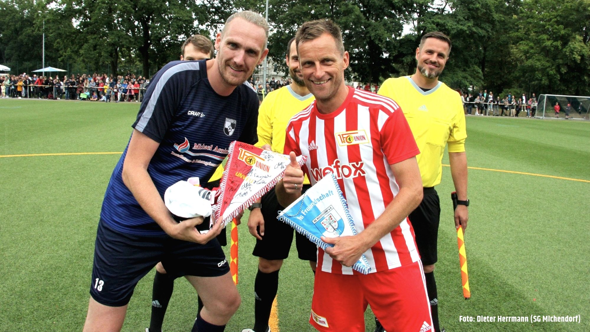 Zwei Fußballspieler lächeln sich an und halten jeweils farbige Trikots in der Hand, während Schiedsrichter im Hintergrund stehen.