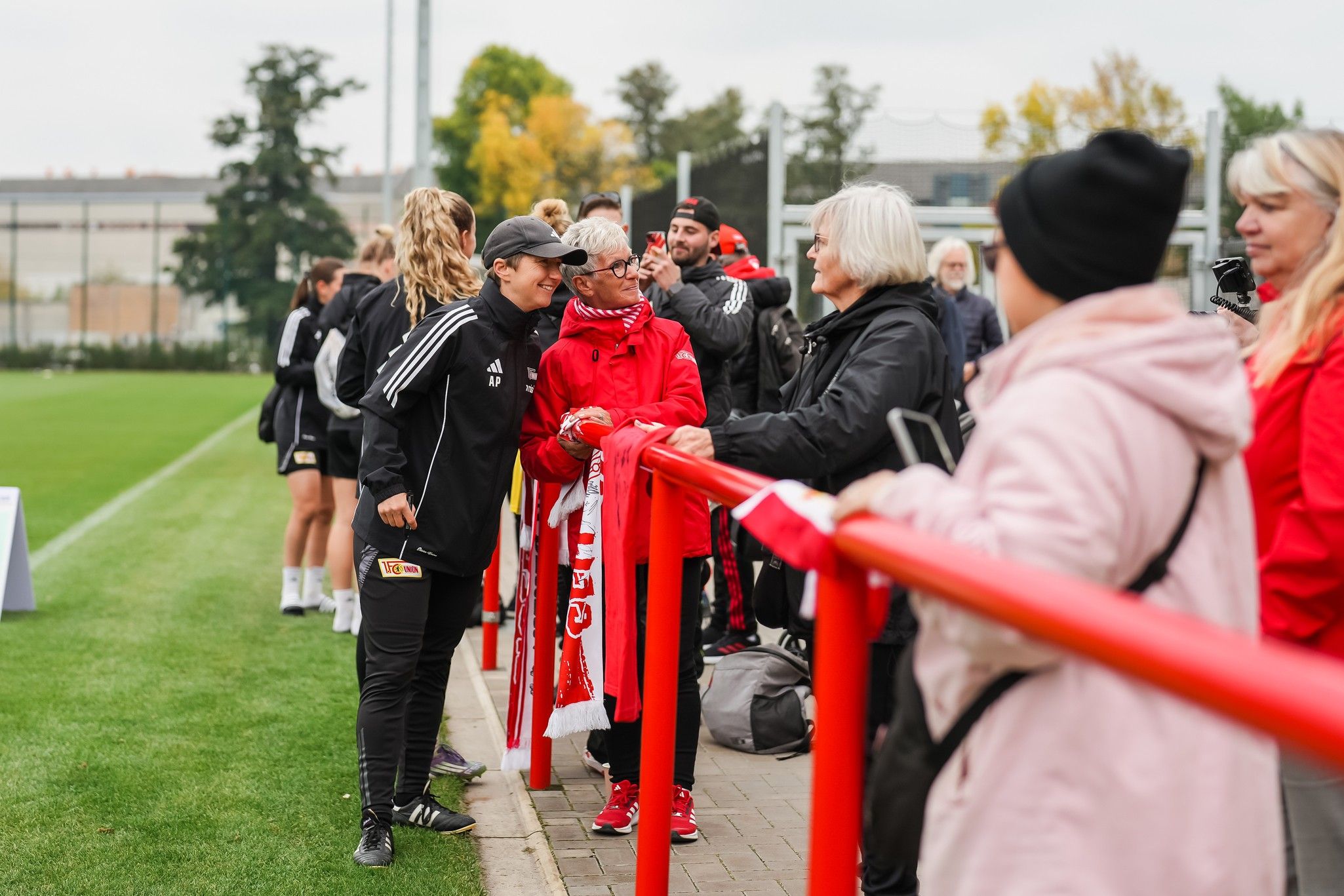 Personen in sportlicher Kleidung stehen an einem roten Zaun, lächeln und unterhalten sich auf einem Fußballfeld. Im Hintergrund sind Bäume.