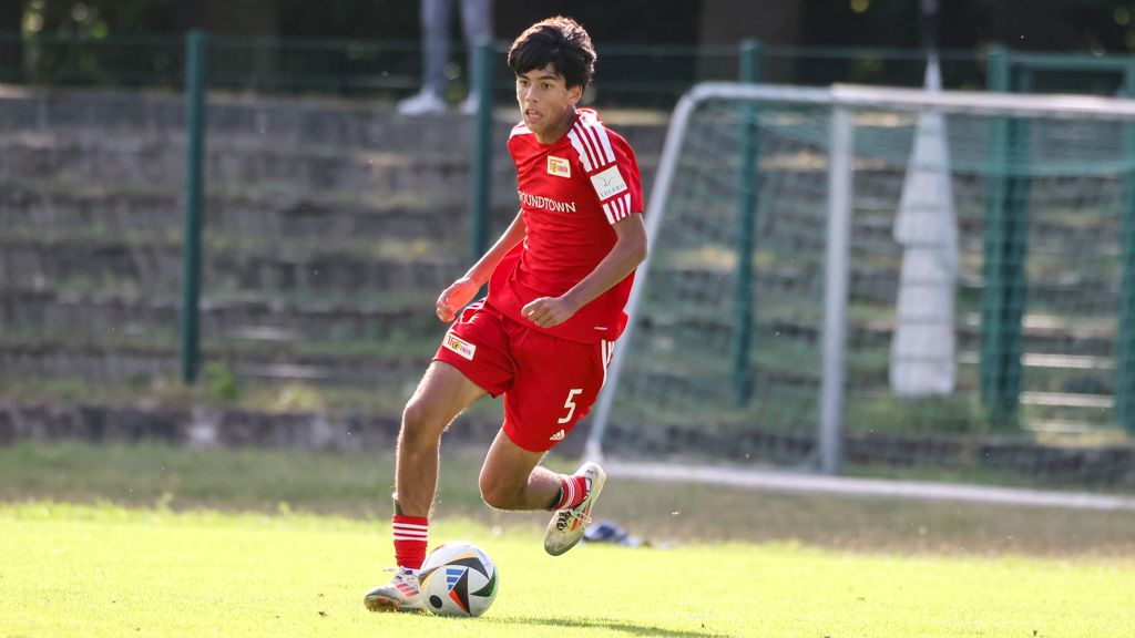 Ein junger Fußballspieler in roter Sportkleidung läuft mit dem Ball auf einem grünen Feld.