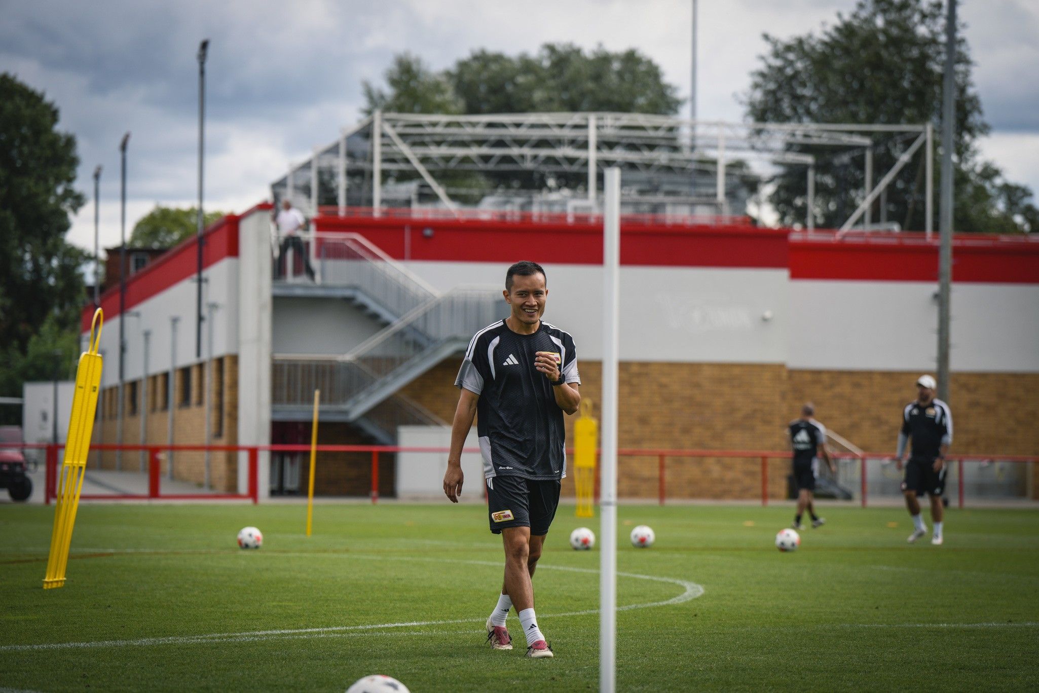 Ein Trainer geht auf einem Fußballfeld mit Trainingsausrüstung, während Spieler im Hintergrund Übungen machen.
