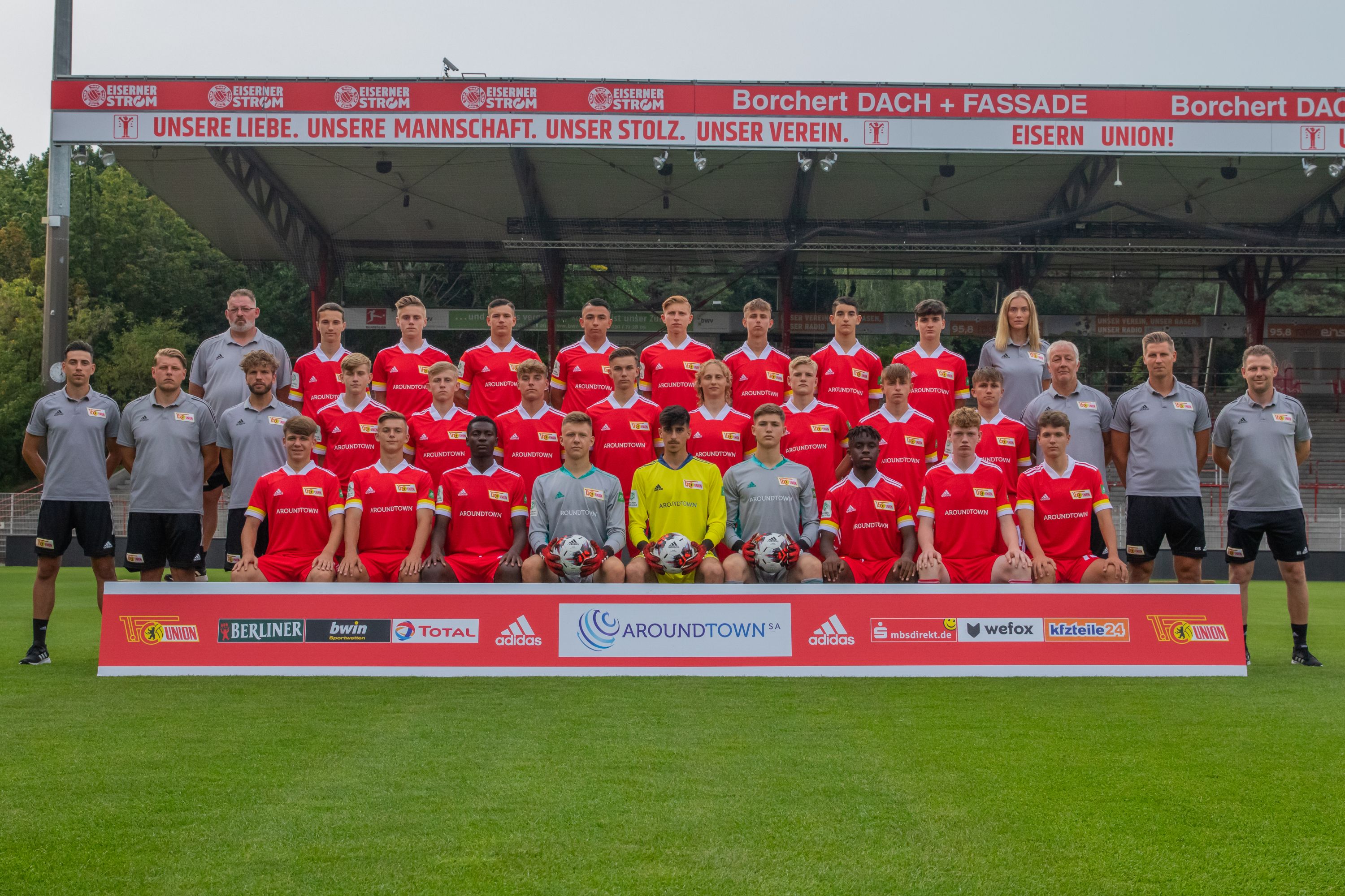 Mannschaftsfoto einer Fußballmannschaft mit Spielern in roten Trikots und Trainern in grauen Shirts auf einem Stadionrasen.