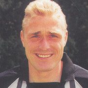 Ein lächelnder Mann mit blonden Haaren trägt ein schwarzes Sporttrikot und steht vor einem unscharfen, grünen Hintergrund.