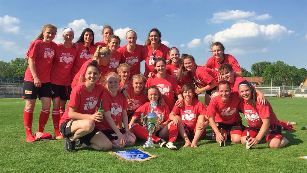 Eine Gruppe von weiblichen Fußballspielerinnen posiert lächelnd im Stadion, umgeben von Trophäen und einem Banner.