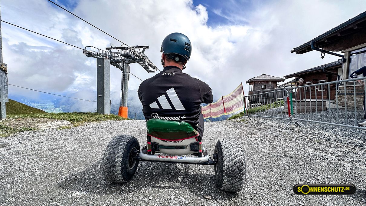 Person sitzt auf einem Dreirad-Bike mit Helm, Blick auf einen Bergweg und eine Seilbahn im Hintergrund.