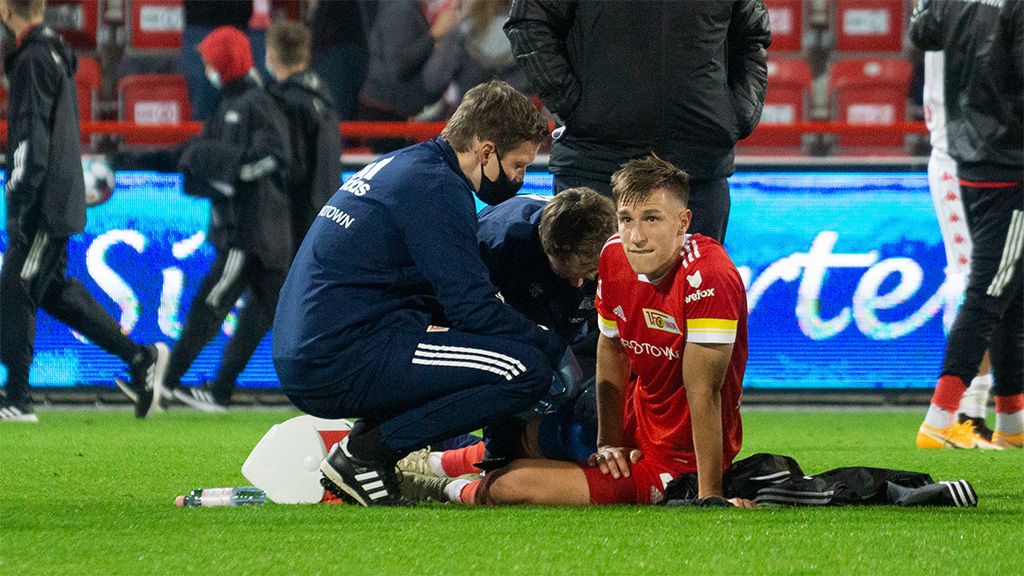 Ein Fußballspieler sitzt auf dem Rasen, während Trainer und Betreuer sich um ihn kümmern.