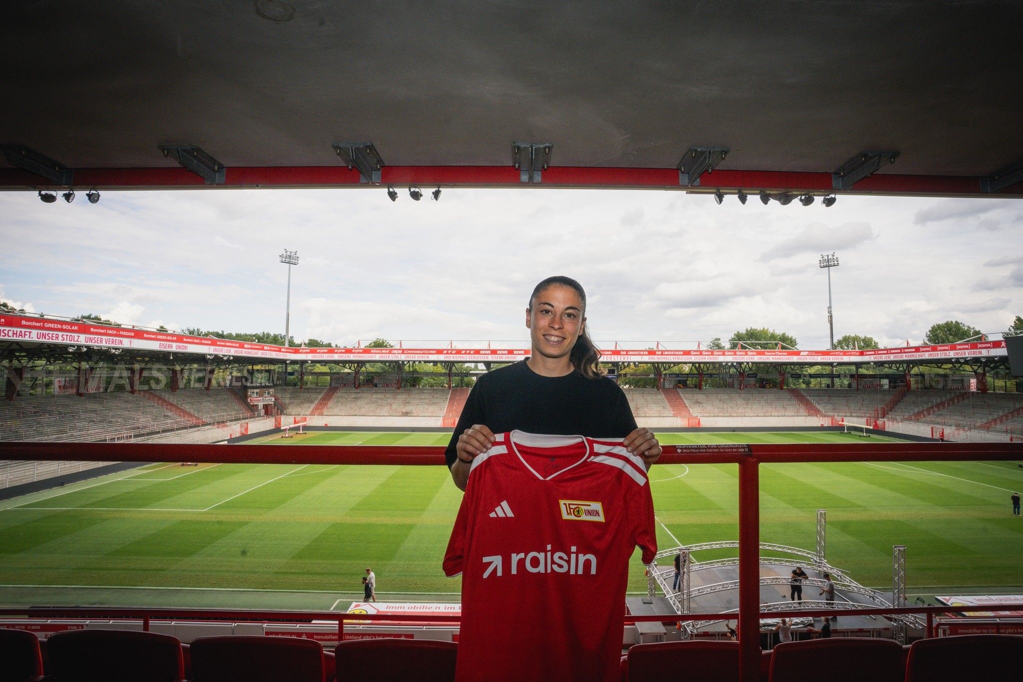 Eine Person hält ein rotes Fußballshirt mit der Aufschrift "raisin" vor einem leeren Stadion mit Rasenfläche.