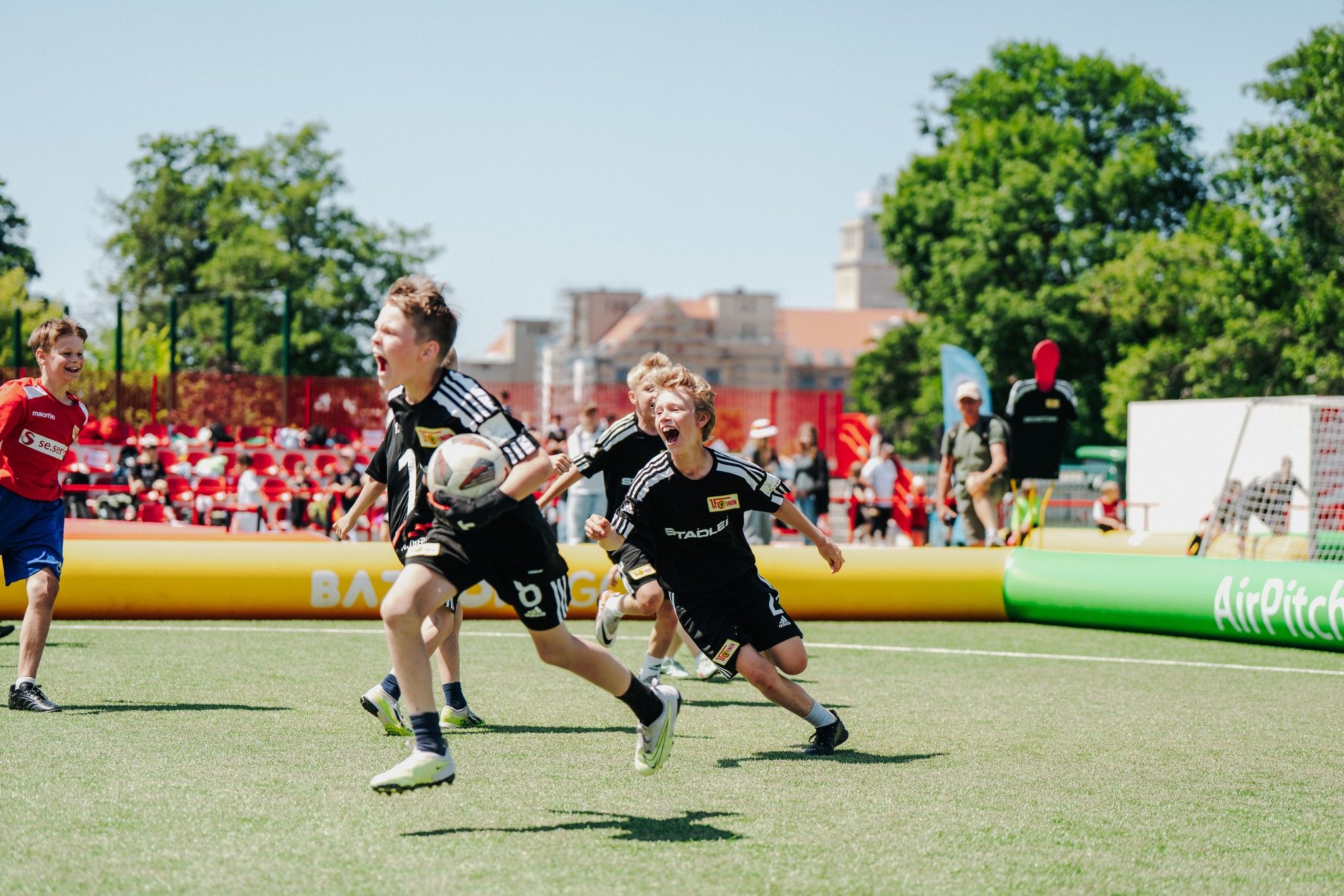 Zwei Kinder in Fußballtrikots rennen auf einem grünen Kunstrasenplatz, ein Ball wird zwischen ihnen gespielt. Zuschauer im Hintergrund.