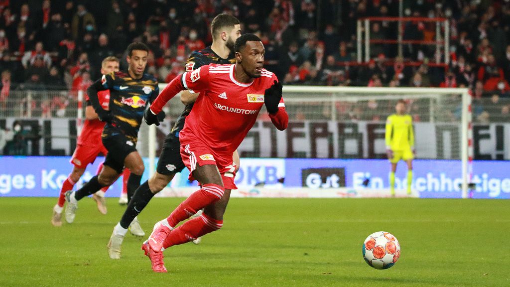Game scene with a Union Berlin player in a red uniform dribbling the ball against a defender from RB Leipzig.