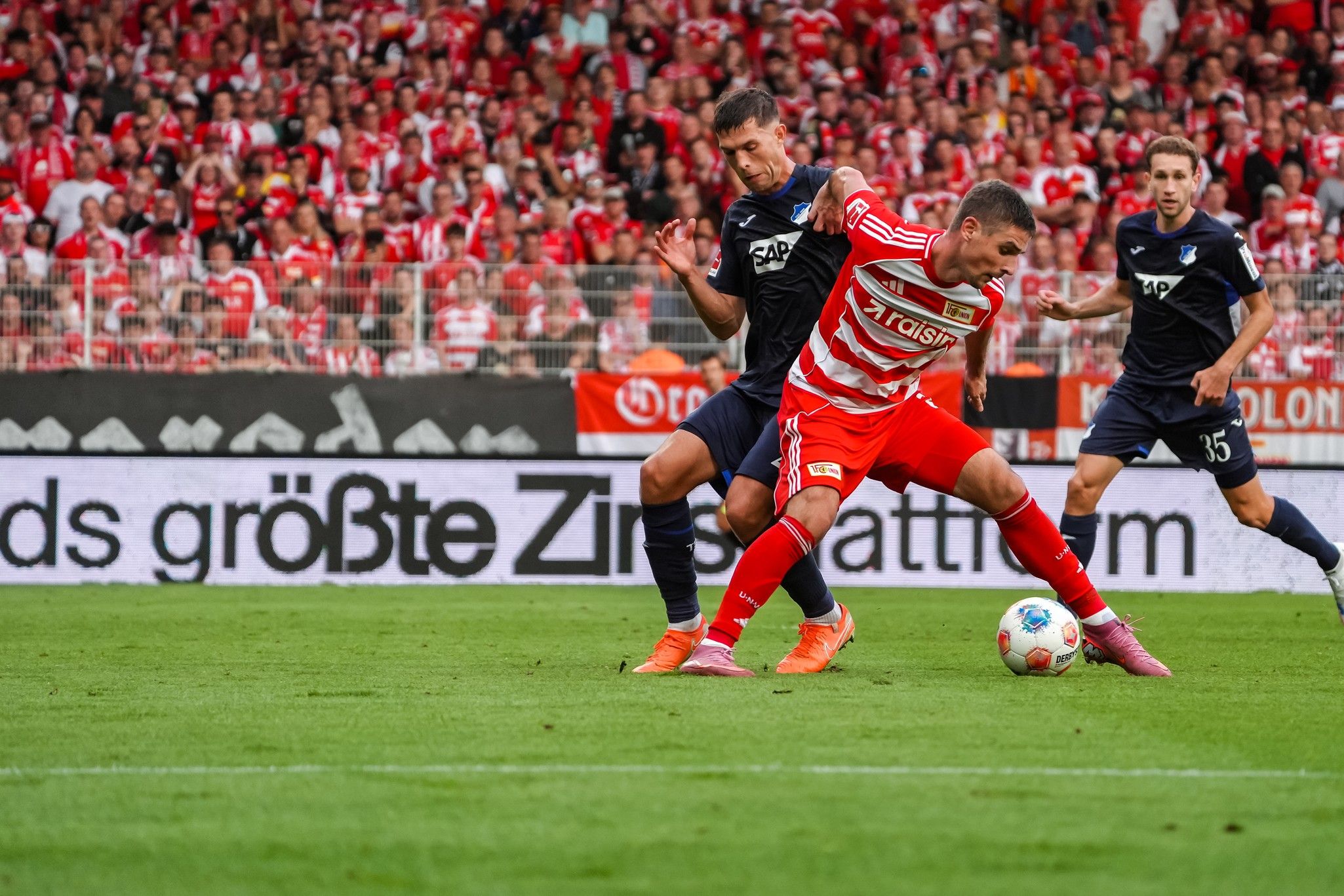 Fußballspiel zwischen zwei Mannschaften, ein Spieler in roten Trikots im Kampf um den Ball gegen einen Spieler in blauen Trikots.