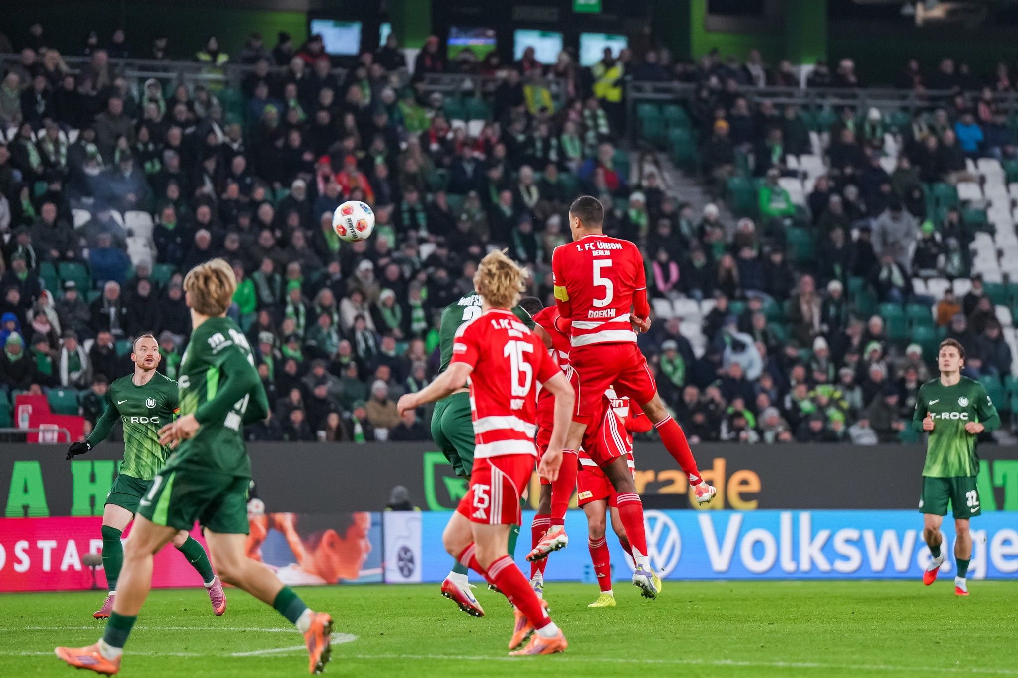 Spielszene im Fußballstadion: Spieler in roten Trikots springen zum Kopfball, Zuschauer im Hintergrund.