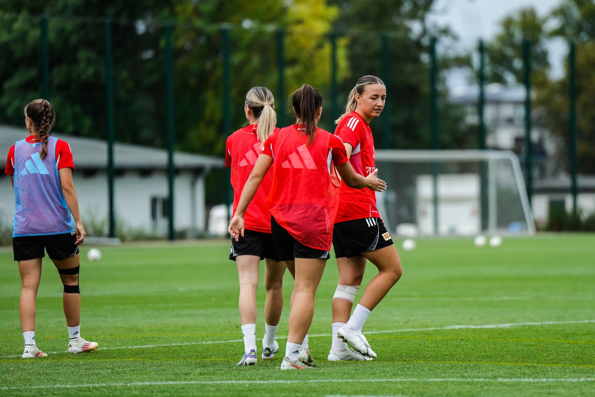 Vier Fußballspielerinnen in roten Trikots trainieren auf einem grünen Platz. Die Umgebung ist ruhig mit Bäumen im Hintergrund.
