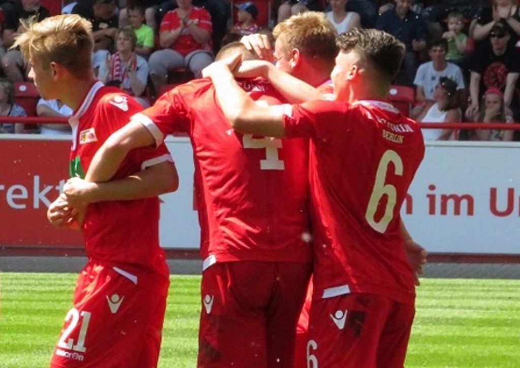 Four football players in red jerseys celebrate a goal on the field.