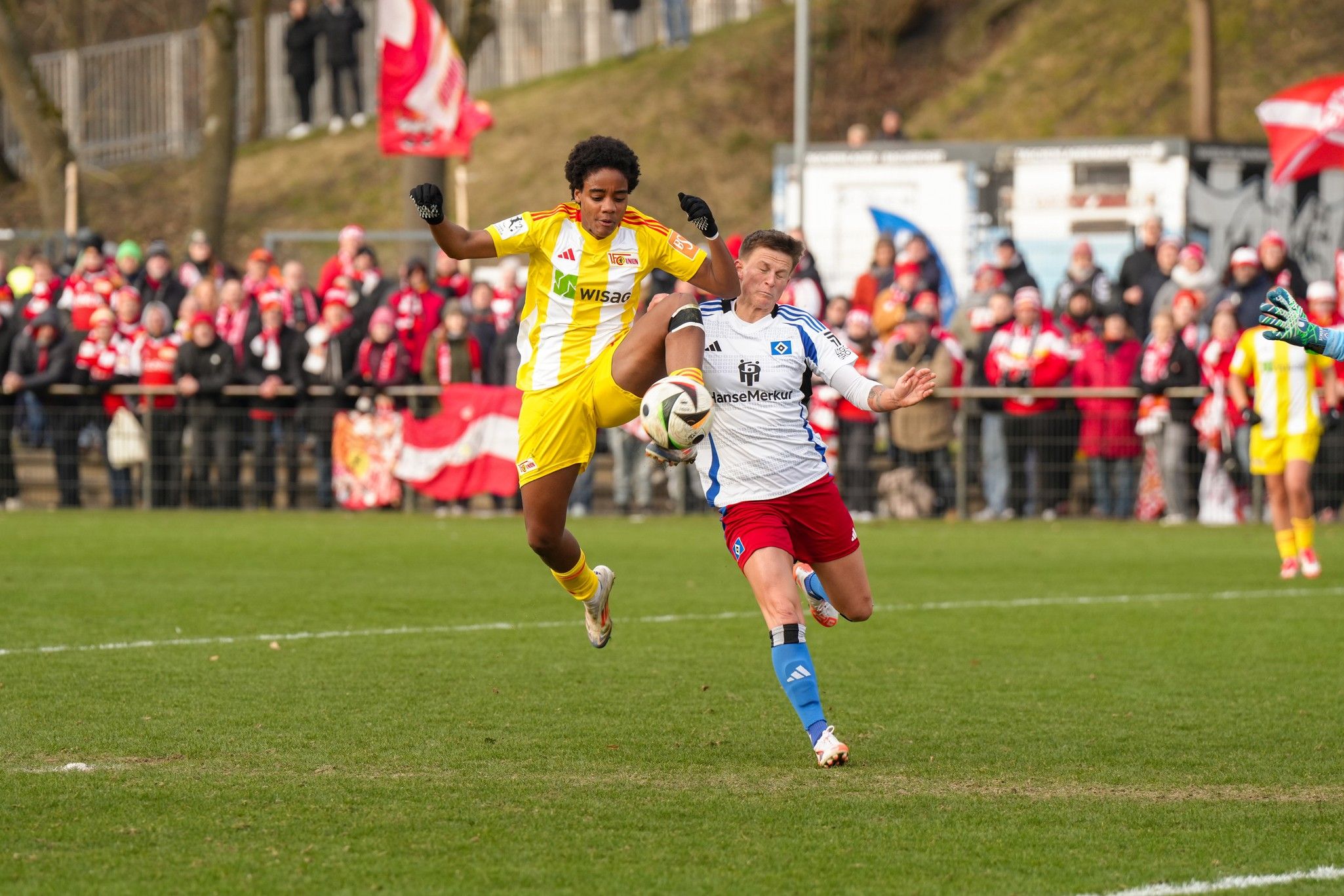 Ein Fußballspieler in gelber Kleidung greift einen Spieler in blauer und weißer Kleidung an, während Fans im Hintergrund jubeln.