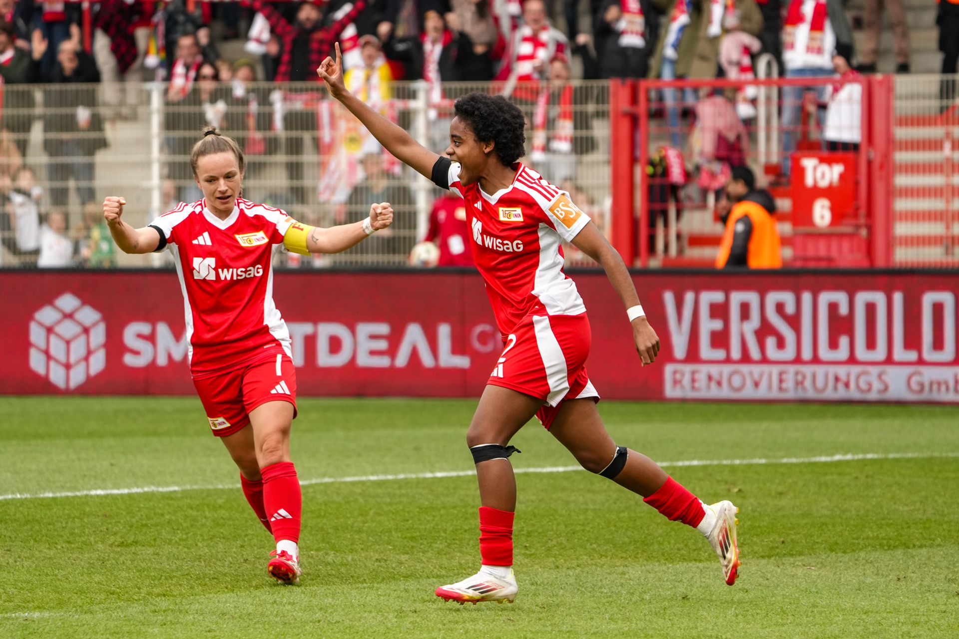 Zwei Spielerinnen in roten Fußballtrikots feiern einen Torerfolg auf dem Spielfeld, im Hintergrund jubelnde Fans.