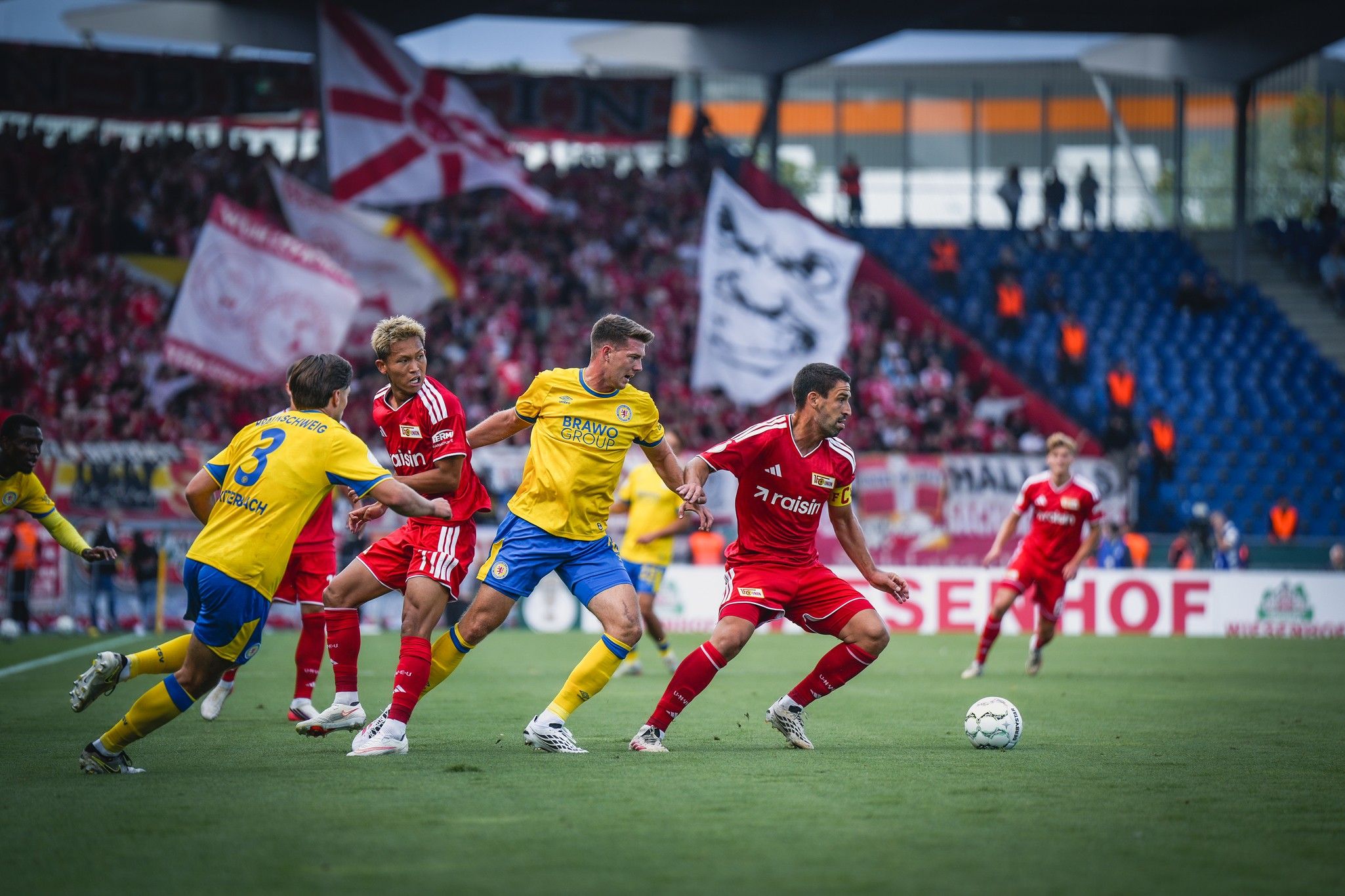 Zwei Fußballteams im Spiel, Spieler in gelben und roten Trikots, leidenschaftliche Fans im Hintergrund mit vielen Vereinsflaggen.