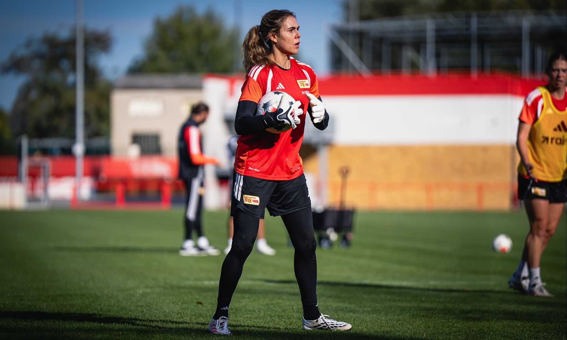Torhüterin in einem rot-schwarzen Trikot beim Training auf dem Fußballfeld, mit zahlreichen anderen Spielern im Hintergrund.