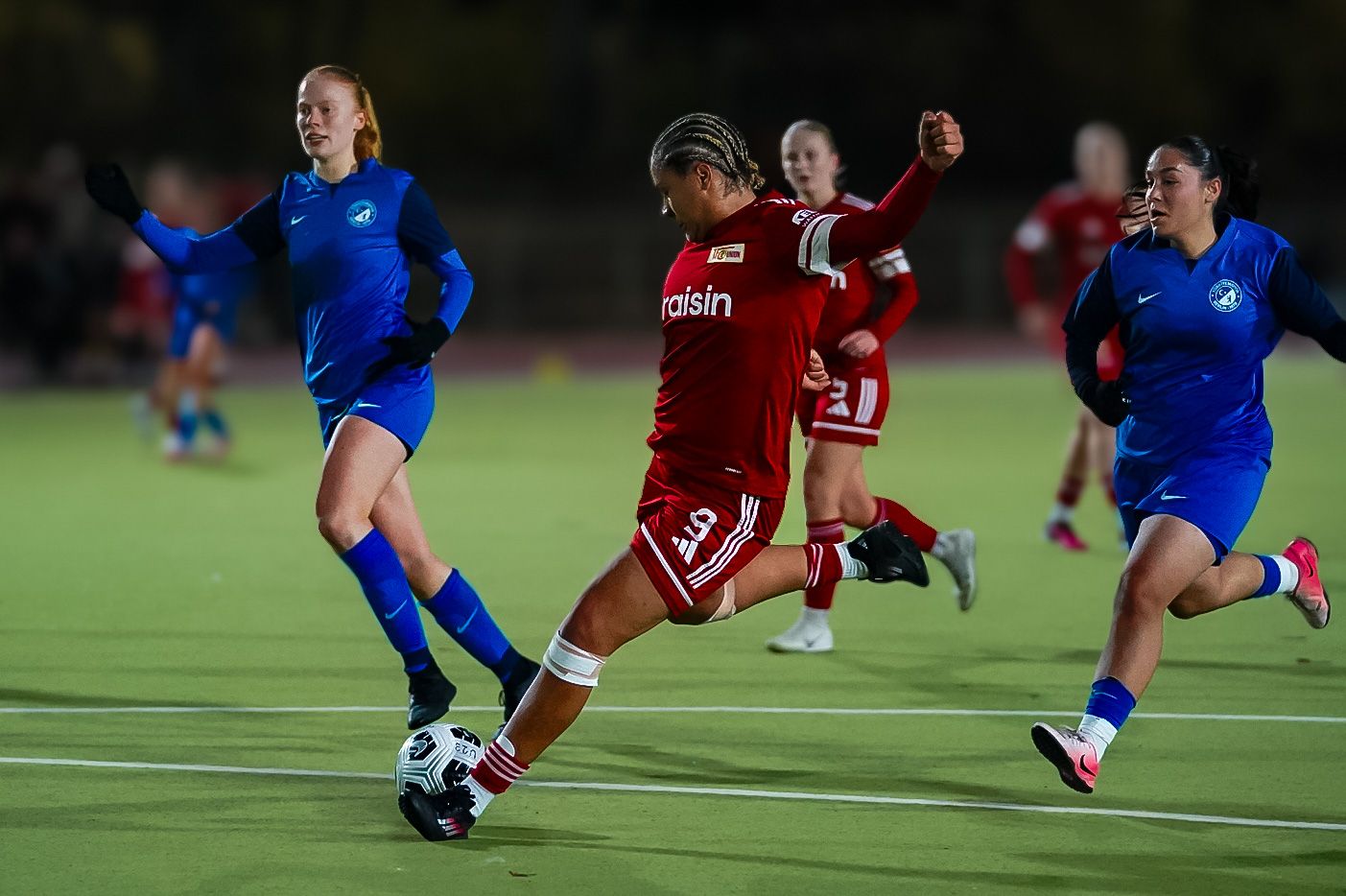 Eine Fußballspielerin in rot schießt den Ball, während zwei Spielerinnen in blau sie verfolgen.