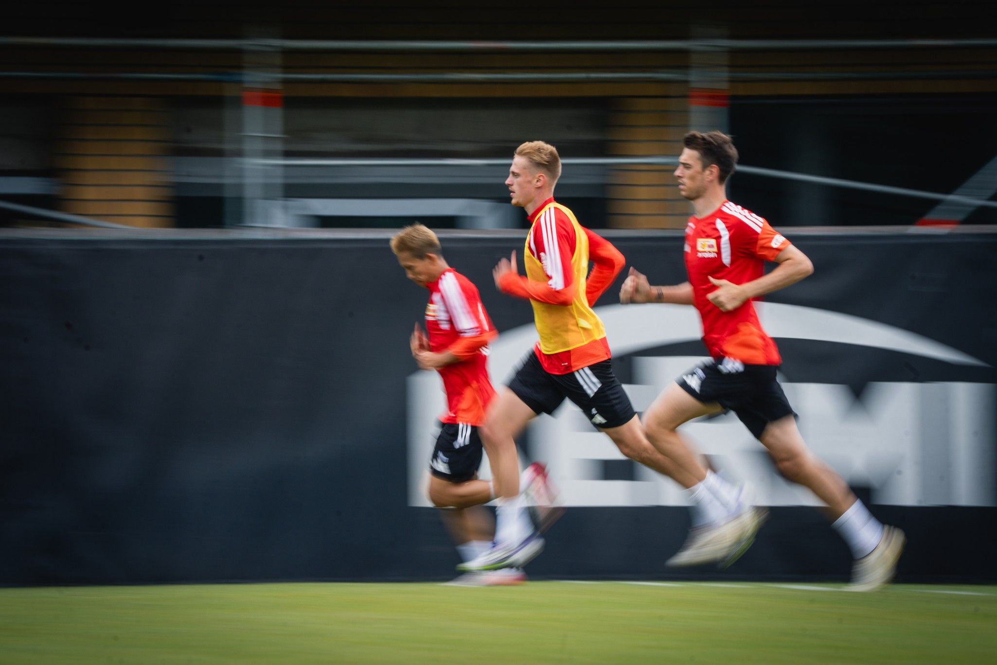 Drei Spieler in roten Trikots sprinten auf einem Fußballfeld, Hintergrund unscharf.