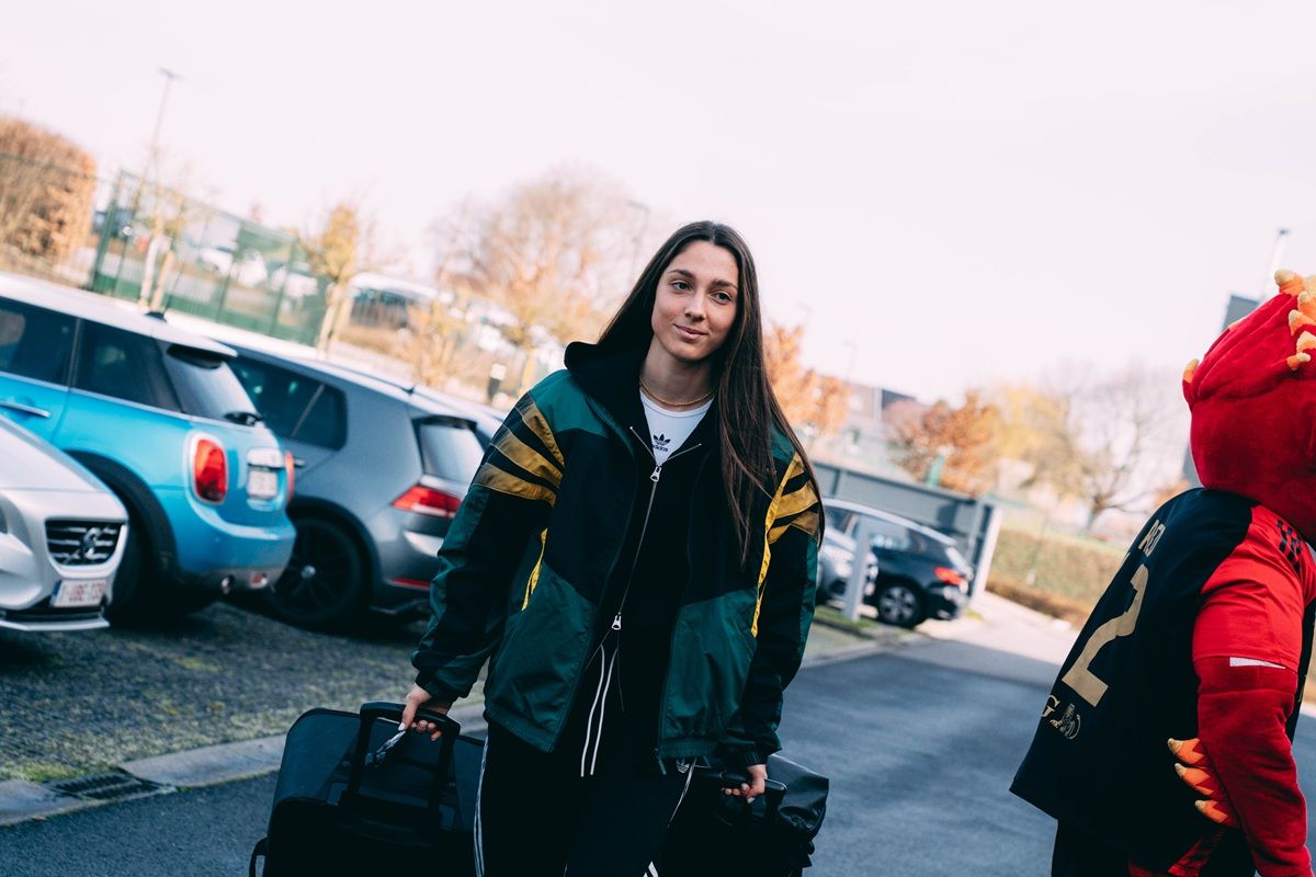 Eine Frau mit langen Haaren trägt zwei schwarze Taschen und geht auf einem Parkplatz vorbei, während ein roter Maskottchen neben ihr steht.