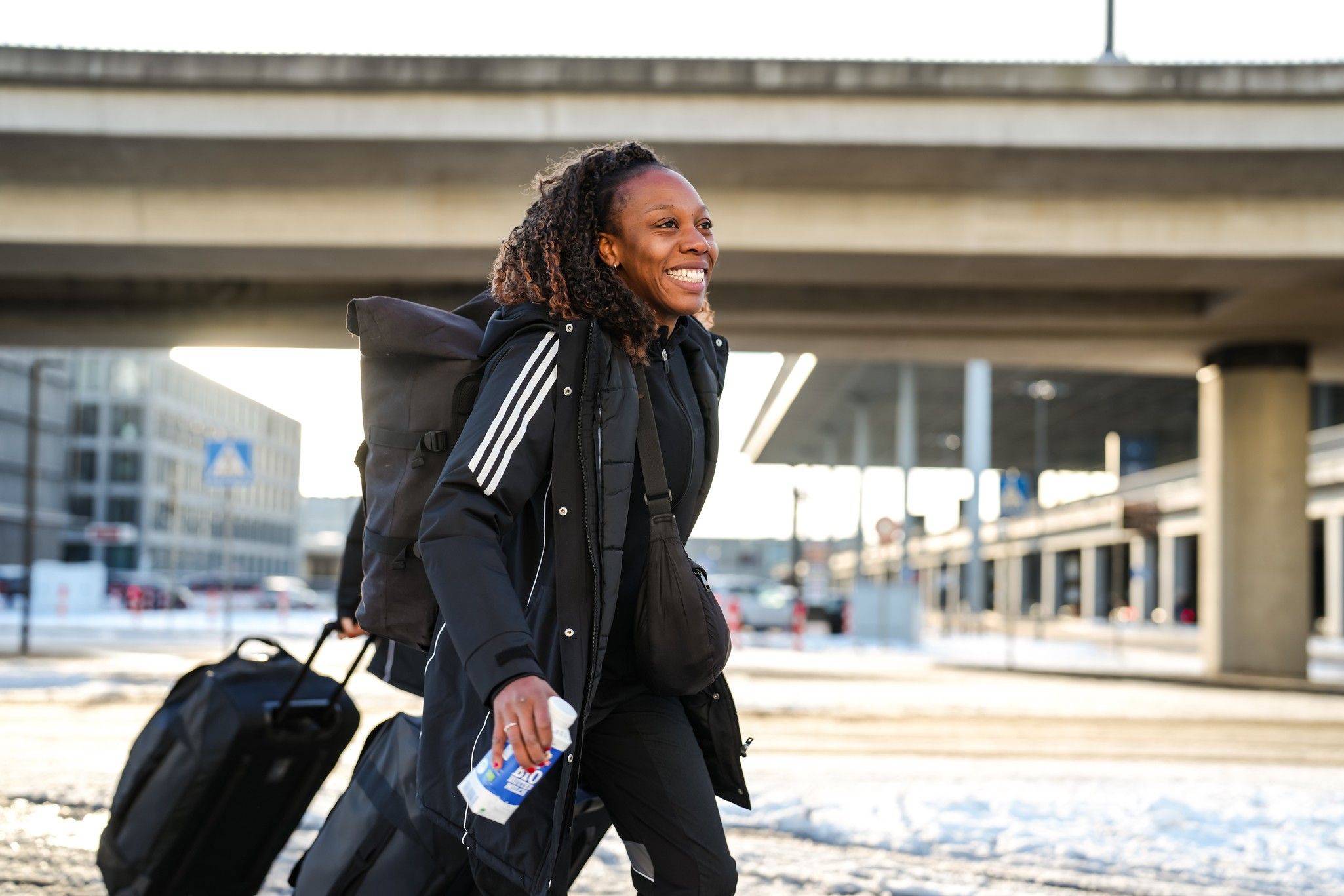Eine Person mit wetterfester Kleidung zieht Gepäck hinter sich und lächelt, während sie auf einem schneebedeckten Gehweg entlanggeht.