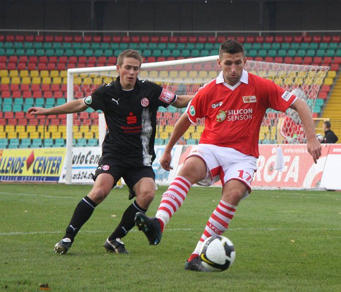 Ein Fußballspiel, in dem ein Spieler mit rotem Trikot den Ball bei einem Dribbling an einem Verteidiger mit schwarzem Trikot vorbeilegt.