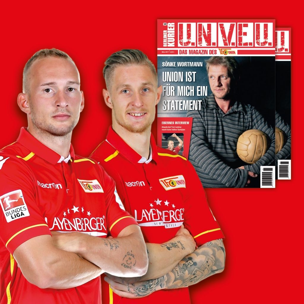 Zwei Fußballspieler in roten Trikots posieren vor einem roten Hintergrund mit einer Zeitschrift im Hintergrund.