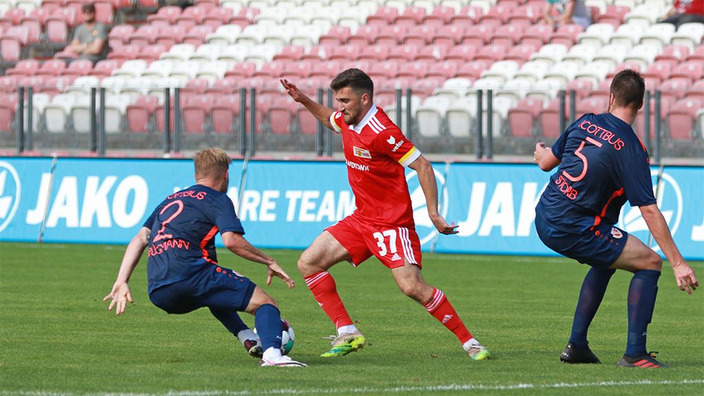 Ein Fußballspieler dribbelt zwischen zwei Gegnern auf einem Sportplatz. Zuschauer sitzen in den Tribünen im Hintergrund.