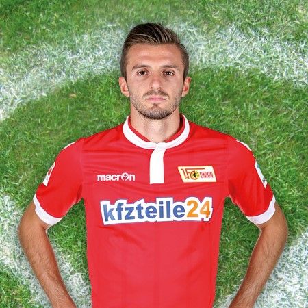 Spieler in rotem Fußballtrikot mit Sponsorenlogo, auf einem Rasenfeld stehend.