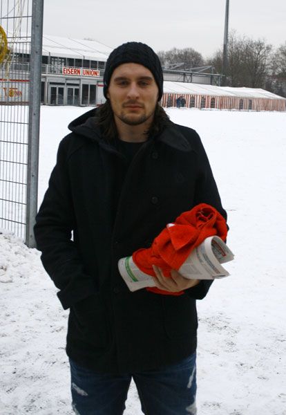Ein Mann mit langen Haaren und schwarzer Jacke steht im Schnee und hält eine rote Decke sowie ein Blatt Papier in den Händen.