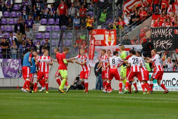 Football players celebrate a goal in front of cheering fans in a stadium.