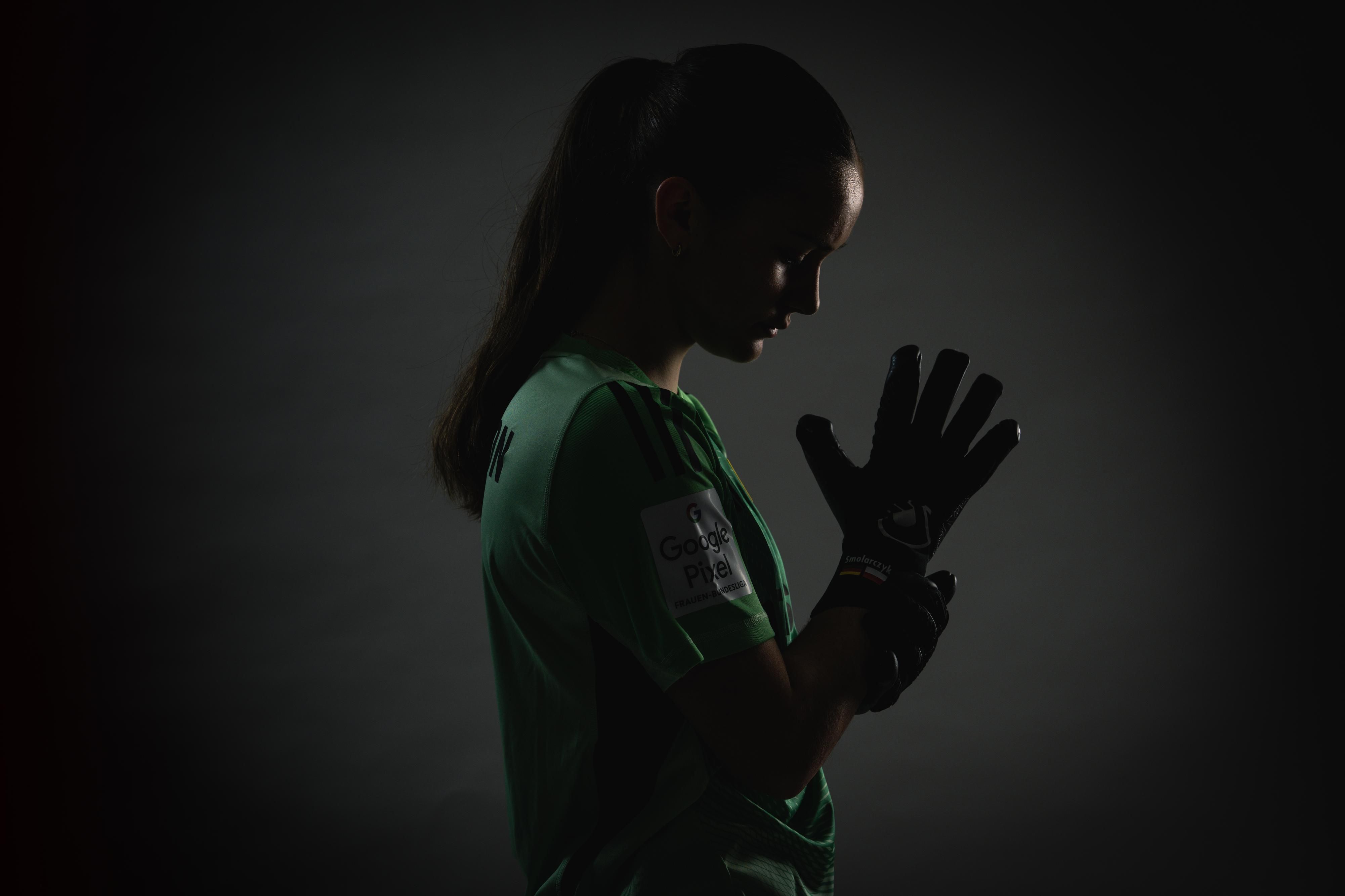 Eine Torhüterin in grüner Sportkleidung und Handschuhen, die nachdenklich in eine dunkle Umgebung blickt.